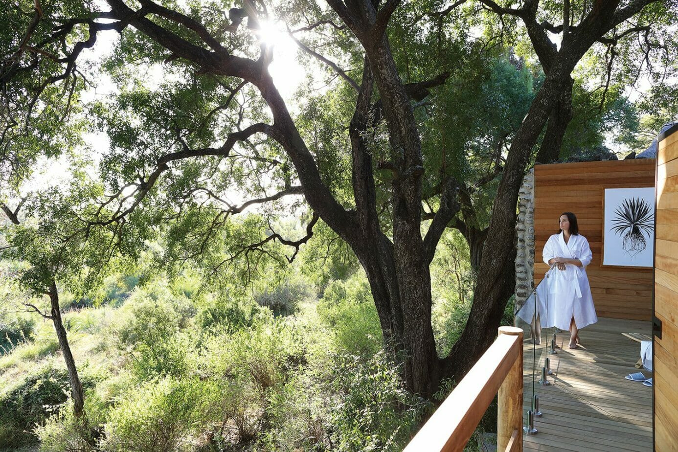 Guest of Londolozi Healing House Retreat standing in white robe on a wooden deck overlooking a canopy of trees.