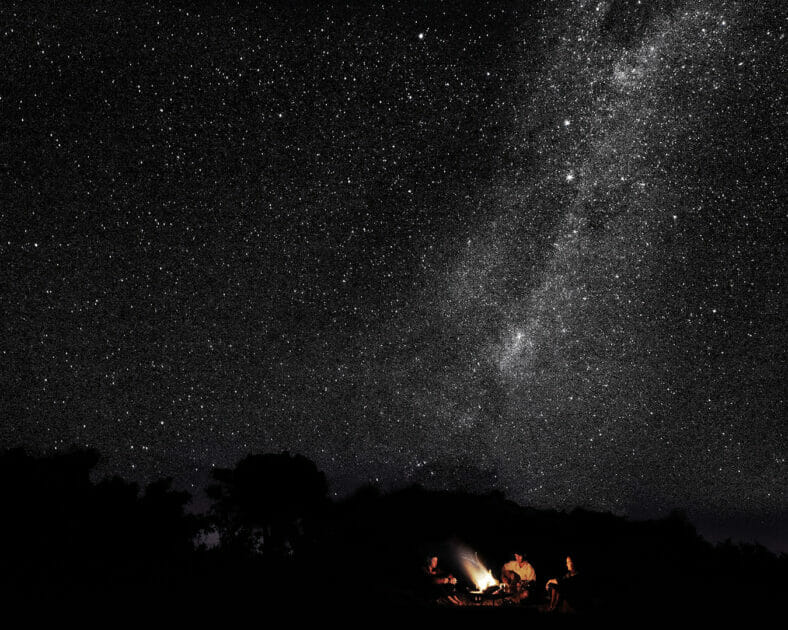 three adults sitting around a remote camp fire with the milky way above them.