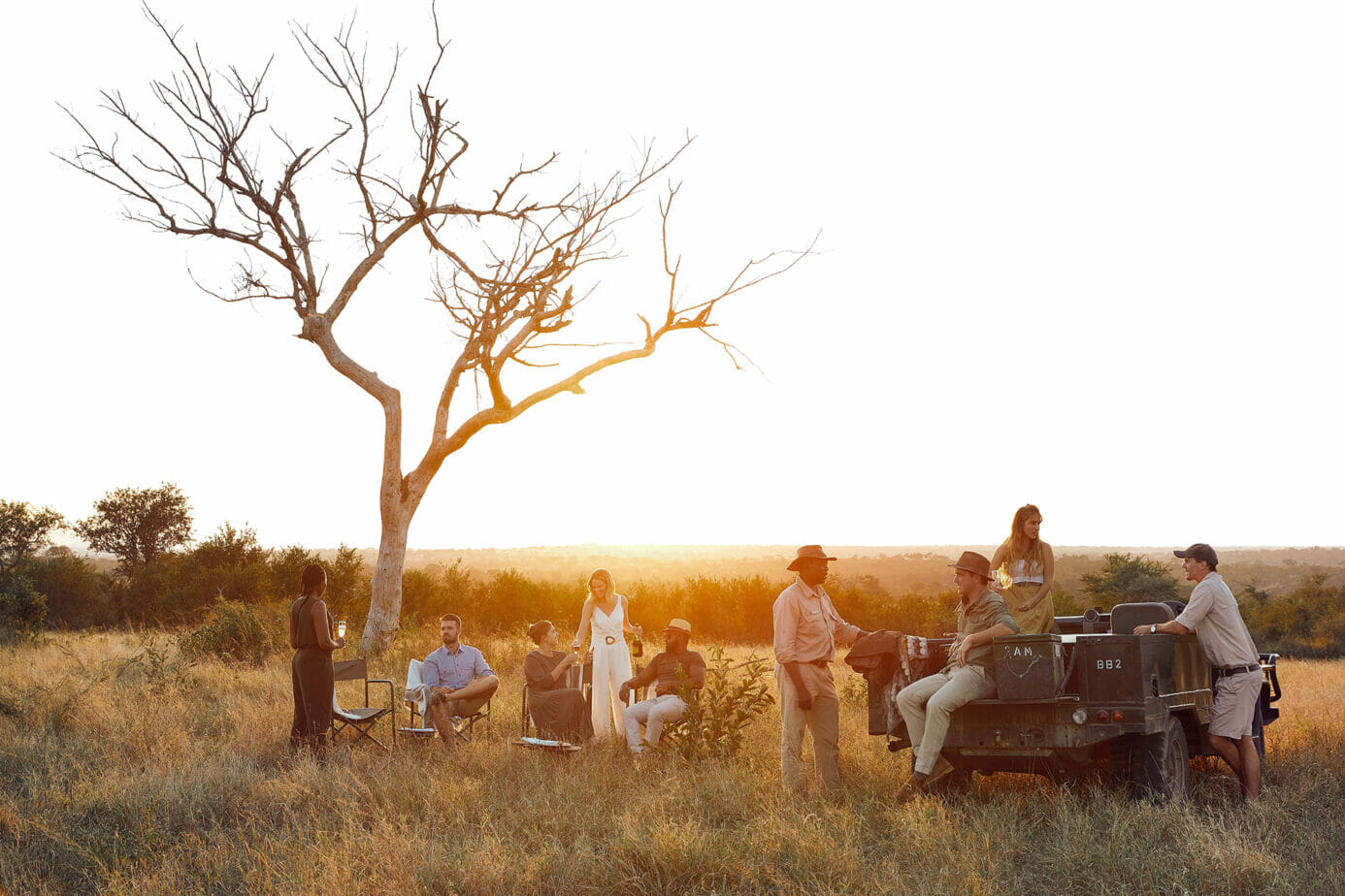 Adult men and women enjoying drinks during sunset in the wilderness at Londolozi, some sitting under a barren tree and others on a green safari vehicle.