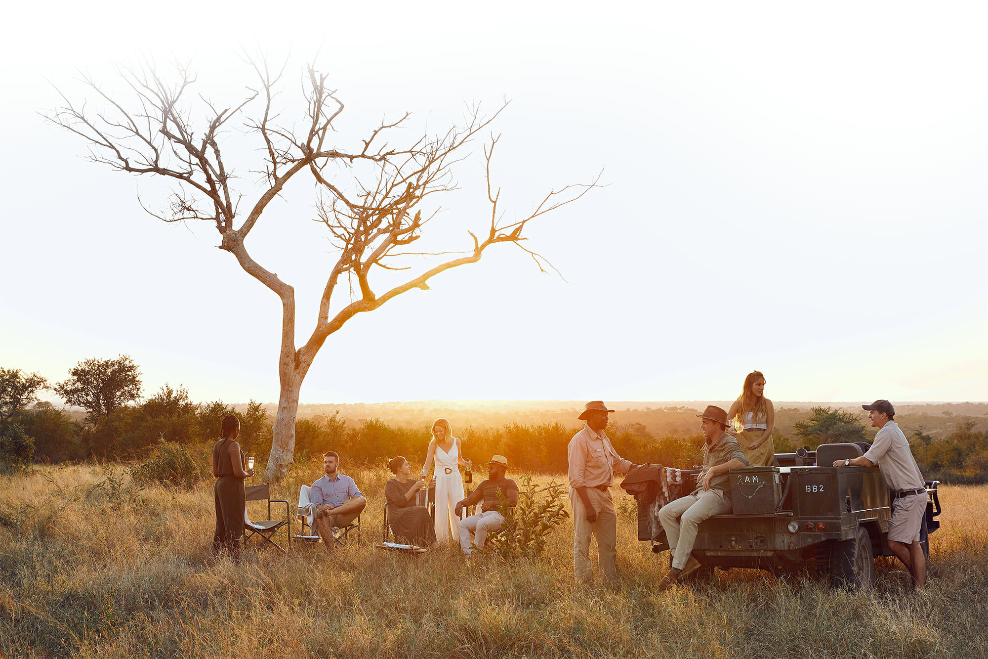 Adult men and women enjoying drinks during sunset in the wilderness at Londolozi, some sitting under a barren tree and others on a green safari vehicle.